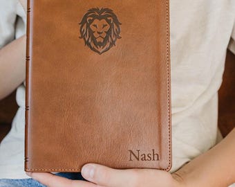 May include: A rectangular brown leather journal featuring a debossed lion head graphic centered on the front cover and the name Nash engraved in the bottom right corner. The journal has visible stitching along the edges and a spine with raised bands.
