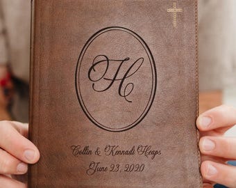 May include: A brown leather-like book cover featuring a centered oval frame with a large script letter H inside. Above the oval is a small cross. Below the oval, the text reads Collin & Kennadi Heaps and the date June 23, 2020, in script font.