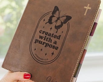 May include: A brown leather-like Bible cover featuring a laser-engraved butterfly, the text "created with a purpose" and "Ephesians 2:10", a small cross symbol, and the name "Victoria" engraved at the bottom, with visible book index tabs.