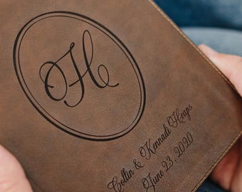May include: A brown leather-like book cover featuring a gold cross at the top, a large script letter H inside an oval, and the engraved text Collin & Kennadi Heaps June 23, 2020. The cover has a smooth, matte finish and dark brown edges.