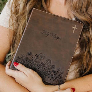 May include: A dark brown leather-like book cover featuring a laser-engraved floral pattern along the bottom edge, the name Lily Harper in script font in the center, and a small cross symbol in the top right corner with visible stitching.