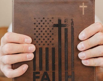 May include: A brown leather-like book cover featuring a laser-engraved design of an American flag combined with a cross. The text FAITH OVER FEAR is printed below the design, with a small cross in the top right and the name M. Carden at the bottom.