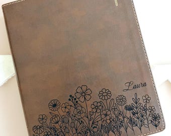 May include: A brown rectangular journal with stitched edges featuring a gold cross icon at the top and a black line art illustration of various wildflowers along the bottom. The name Laura is engraved in black script text above the floral design.