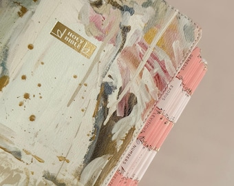 May include: A white and gold Holy Bible with a painted floral design. The cover features a gold rectangular emblem with the words "Holy Bible". Pink and white page dividers are visible, with book names printed on them.