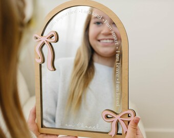 May include: An arched wooden mirror featuring two pink decorative bows attached to the frame. The glass is etched with the phrases I am important, I am kind, I am smart, I am brave, I am loved, and the name I am Chanel written in white script.