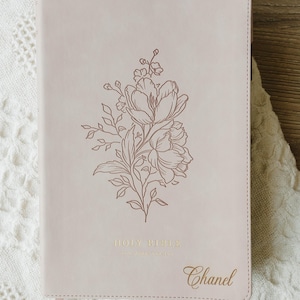 Puede incluir: Una Biblia de color rosa claro con una cubierta lisa que presenta una ilustración floral de líneas marrón oscuro centrada en la parte frontal. Las palabras HOLY BIBLE están impresas en lámina dorada cerca de la base, con el nombre Chanel grabado.