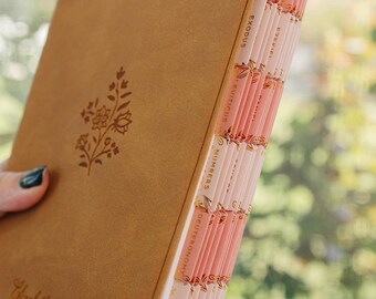 May include: A tan leather-bound book featuring a debossed floral design on the front cover and the name Elizabeth Grace in script. The page edges are indexed with pink tabs labeled with book names including Exodus, Leviticus, Numbers, and Joshua.