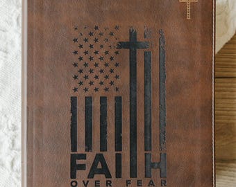 May include: A brown faux leather journal with a black embossed design featuring an American flag integrated with a cross, the text FAITH OVER FEAR, a small gold cross in the top right corner, and the personalized name M. Cardon in the bottom right.