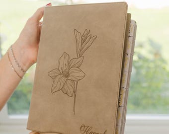 May include: A tan-colored notebook with a smooth, matte cover featuring a dark brown line-art illustration of a lily flower. The name Hannah is engraved in a cursive script in the bottom right corner. The notebook has visible tabbed dividers.