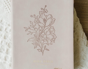 May include: A light pink Bible with a smooth cover featuring a dark brown line art floral illustration centered on the front. The words HOLY BIBLE are printed in gold foil near the bottom, with the name Chanel embossed in gold script below it.