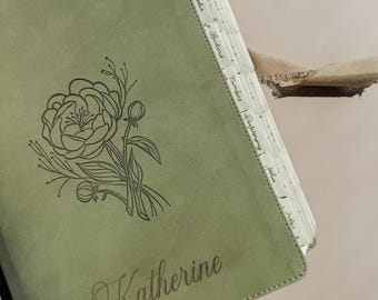 May include: A sage green journal with a matte finish featuring a dark green line art illustration of a peony flower and the name Katherine engraved on the front cover. The journal includes white tabbed dividers with printed book names.