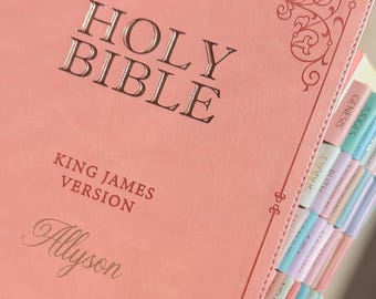 May include: A coral-colored Holy Bible, King James Version, with the name Allyson embossed on the front cover. The cover features decorative scrollwork at the corners. The book includes a series of pastel-colored index tabs along the side edge.