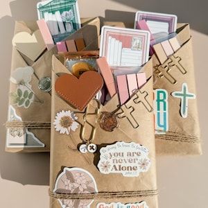 May include: Three brown paper wrapped packages decorated with stickers, including a brown heart, cross-shaped paper clips, floral designs, and text reading "you are never Alone" and "God is good." Each package contains stationery items.