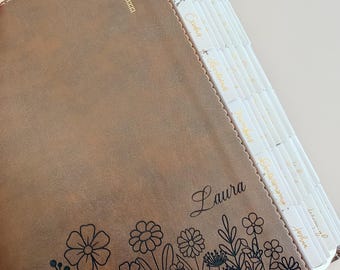 May include: A brown leather-like book cover featuring a gold cross emblem at the top, the name Laura in cursive script, and a black line-art floral pattern along the bottom edge. The book includes white index tabs with gold text for book names.