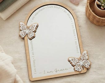 May include: An arched mirror with a light wood frame featuring two white laser-cut butterfly accents. The mirror surface is printed with the text: I am important, I am kind, I am smart, I am brave, I am loved, I am strong, I am beautiful, and I am Grace.