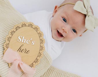 May include: A baby wearing a white onesie and a light green bow headband, wrapped in a cream-colored knit blanket. An oval wooden plaque with a scalloped edge features the text "She's here" and is decorated with a soft pink ribbon.