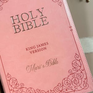 May include: A light pink Bible with a smooth cover featuring dark pink decorative scrollwork in the corners. The text reads HOLY BIBLE, KING JAMES VERSION, and Mari's Bible in a script font. The cover has a stitched edge and a light ribbon.