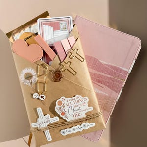 May include: A brown paper envelope containing stationery items, including a pink heart, cross-shaped paper clips, a wax seal, and stickers. One sticker reads, I can do ALL THINGS through Christ who strengthens me. A pink notebook is tucked behind the envelope.