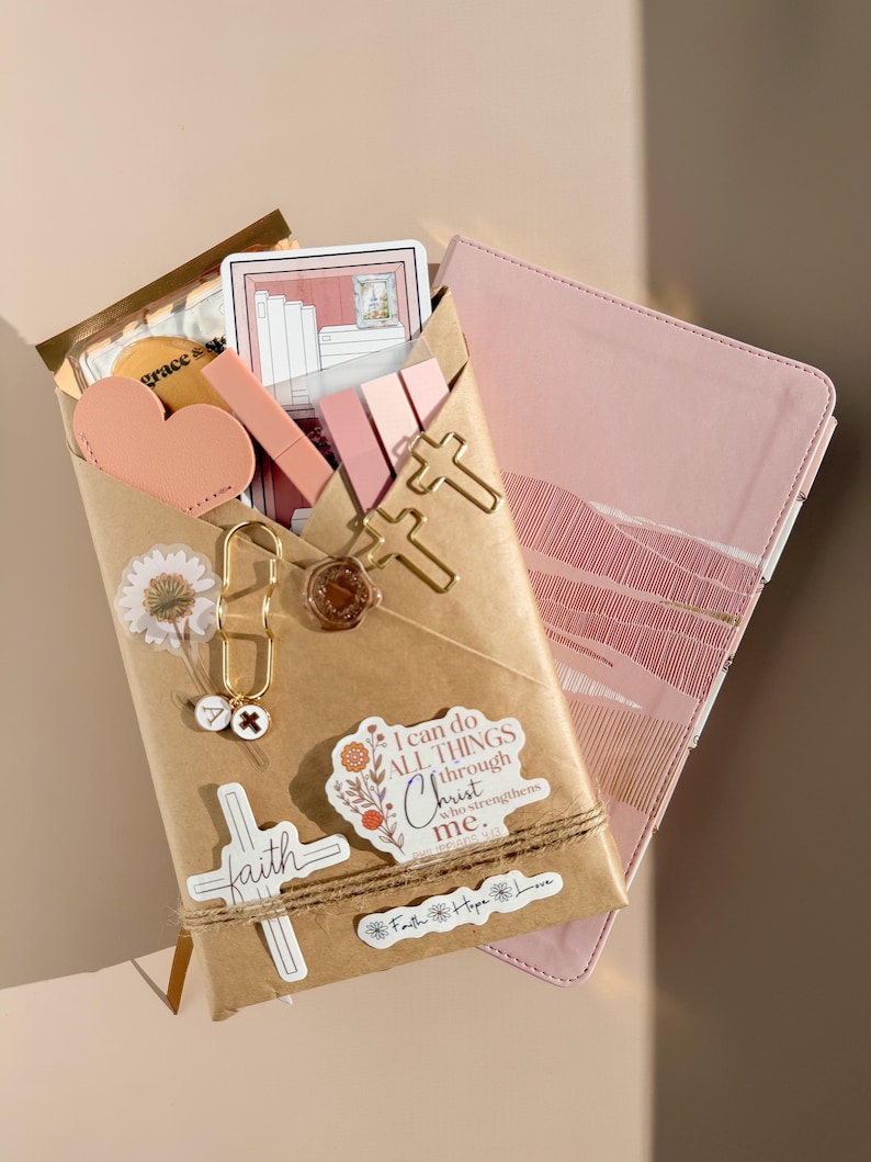 May include: A brown paper envelope containing stationery items, including a pink heart, cross-shaped paper clips, a wax seal, and stickers. One sticker reads, I can do ALL THINGS through Christ who strengthens me. A pink notebook is tucked behind the envelope.