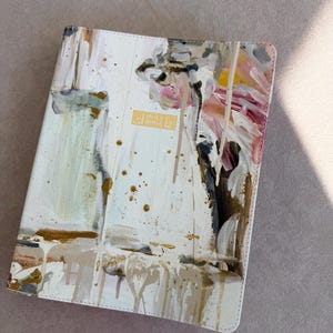 May include: A white Bible cover with an abstract painted design in shades of beige, gold, pink, and black. The cover has a gold rectangular label that reads "HOLY BIBLE". The design has a textured, dripped paint effect.