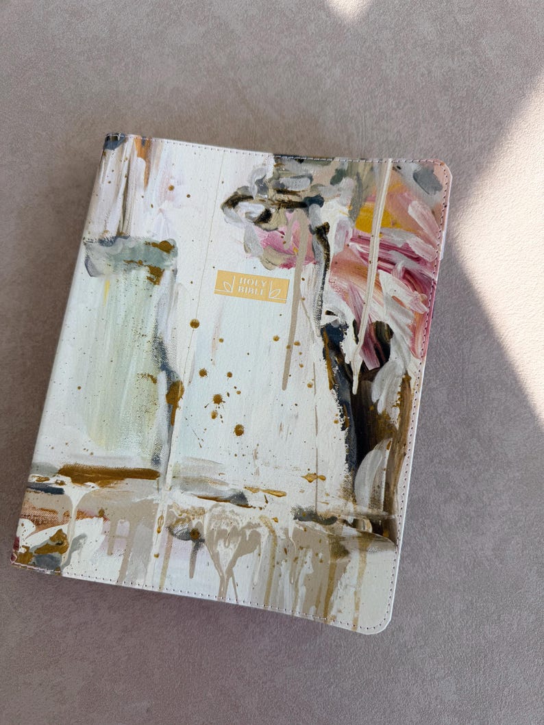 May include: A white Bible cover with an abstract painted design in shades of beige, gold, pink, and black. The cover has a gold rectangular label that reads "HOLY BIBLE". The design has a textured, dripped paint effect.