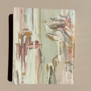 May include: A rectangular notebook with a textured cover featuring an abstract painted design in shades of sage green, soft pink, white, and metallic gold. The paint application includes vertical brushstrokes and splatters across the surface.