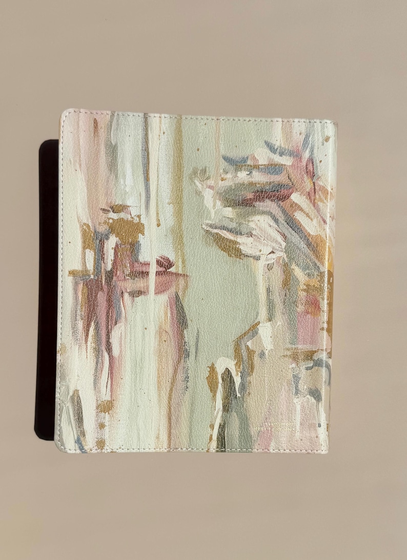 May include: A rectangular notebook with a textured cover featuring an abstract painted design in shades of sage green, soft pink, white, and metallic gold. The paint application includes vertical brushstrokes and splatters across the surface.