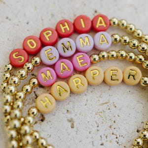 May include: Gold beaded bracelets with names spelled out in colorful letter beads. The names Sophia, Emma, and Harper are visible. The letter beads are in shades of red, pink, and peach.