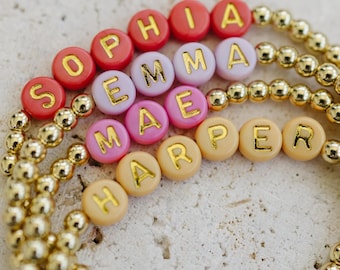 May include: Four stretch bracelets featuring metallic gold-tone beads and round plastic beads in shades of red, light purple, pink, and beige. Each bracelet displays a name in gold lettering: SOPHIA, EMMA, MAE, and HARPER, arranged in a stacked formation.