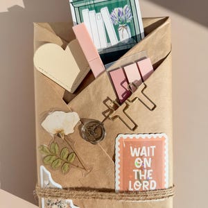 May include: A brown paper package decorated with a heart-shaped tag, a pink pen, sticky notes, gold paper clips, a wax seal, a dried flower sticker, and a rectangular sticker with the text WAIT ON THE LORD, all tied with twine.