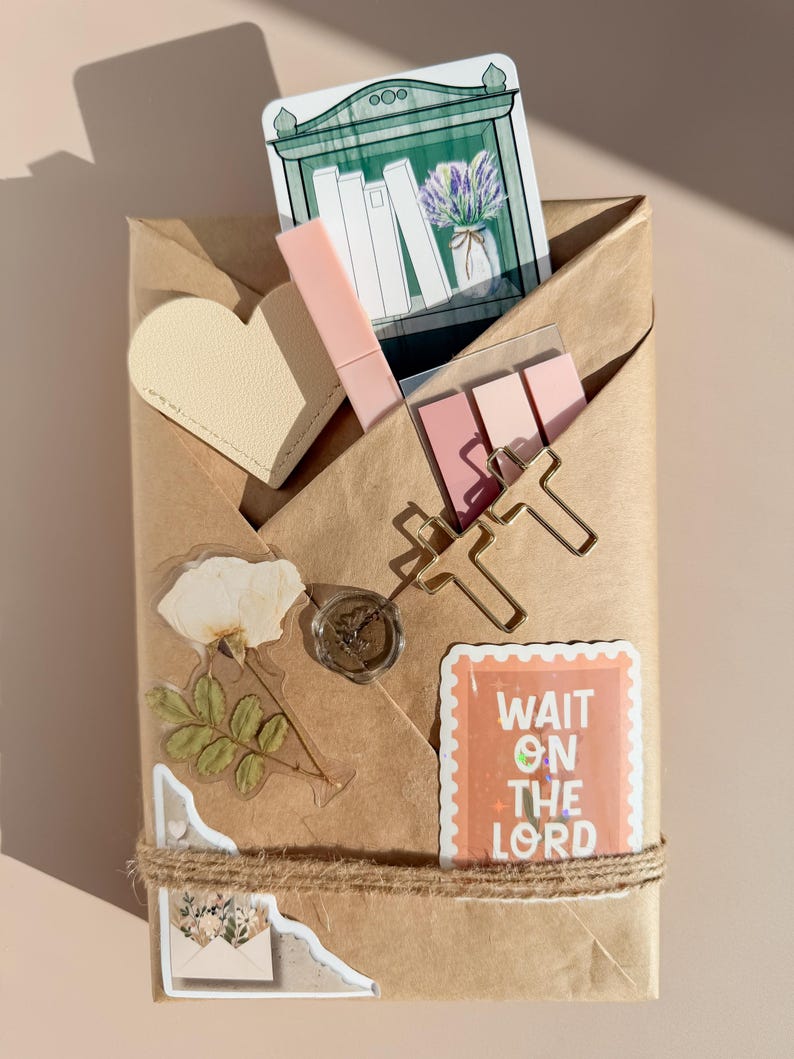 May include: A brown paper package decorated with a heart-shaped tag, a pink pen, sticky notes, gold paper clips, a wax seal, a dried flower sticker, and a rectangular sticker with the text WAIT ON THE LORD, all tied with twine.