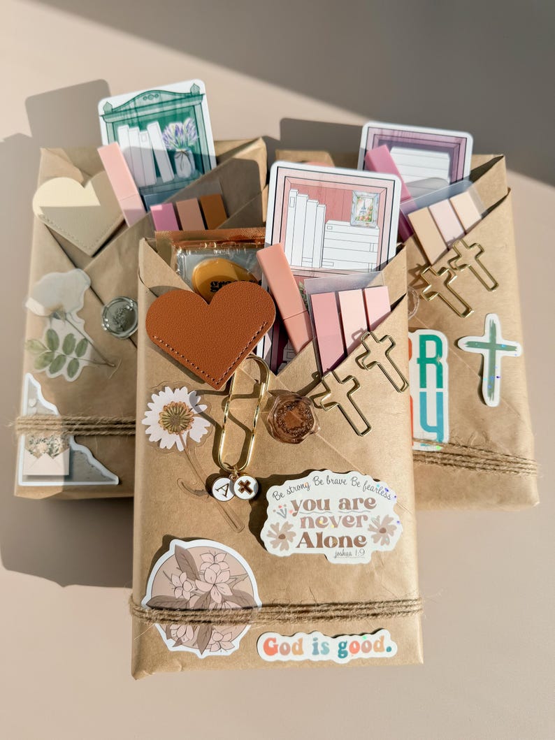May include: Three brown paper wrapped packages decorated with stickers, including a brown heart, cross-shaped paper clips, floral designs, and text reading "you are never Alone" and "God is good." Each package contains stationery items.