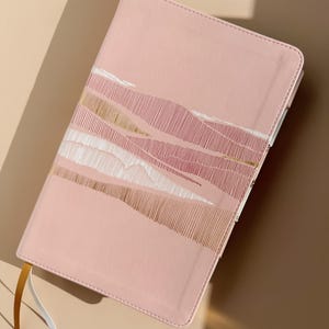May include: A light pink notebook with a rectangular shape and rounded corners. The cover features a decorative pattern of horizontal lines in white and metallic gold. Two thin ribbon bookmarks, one gold and one white, extend from the bottom edge.