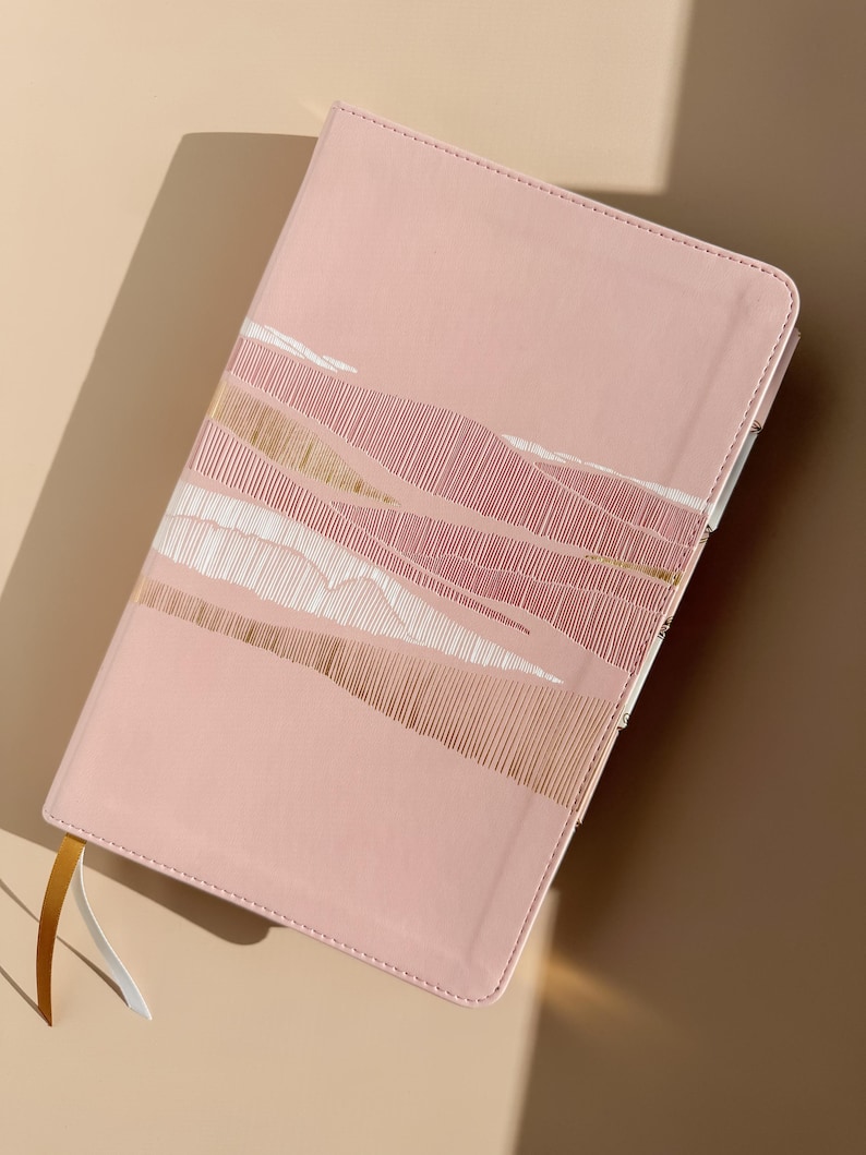May include: A light pink notebook with a rectangular shape and rounded corners. The cover features a decorative pattern of horizontal lines in white and metallic gold. Two thin ribbon bookmarks, one gold and one white, extend from the bottom edge.