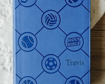 May include: A blue book cover featuring a diamond grid pattern with embossed circular icons of a hockey stick, football, volleyball, tennis ball, soccer ball, basketball, baseball, and the text Holy Bible and the name Travis embossed on the front.