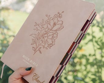 May include: A light pink Holy Bible King James Version with a floral line art illustration on the cover. The name Chanel is embossed at the bottom. The book features dark red and black index tabs along the side for navigating the various books.