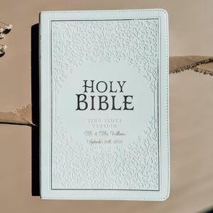 May include: A white Holy Bible with a debossed floral vine pattern on the cover. The text reads HOLY BIBLE, KING JAMES VERSION, Mr. & Mrs. Williams, and September 26th, 2026. The cover features a rectangular border and stitched edges.