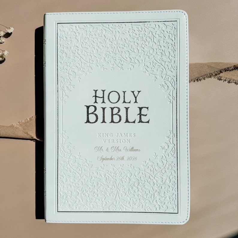 May include: A white Holy Bible with a debossed floral vine pattern on the cover. The text reads HOLY BIBLE, KING JAMES VERSION, Mr. & Mrs. Williams, and September 26th, 2026. The cover features a rectangular border and stitched edges.