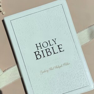 May include: A white Holy Bible with a textured floral embossed border and silver-toned edges. The cover features the text HOLY BIBLE, KING JAMES VERSION, and the personalized names Zachery And Aaliyah Pilcher printed in a dark serif font.