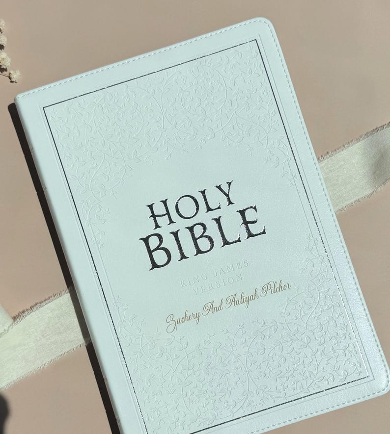 May include: A white Holy Bible with a textured floral embossed border and silver-toned edges. The cover features the text HOLY BIBLE, KING JAMES VERSION, and the personalized names Zachery And Aaliyah Pilcher printed in a dark serif font.