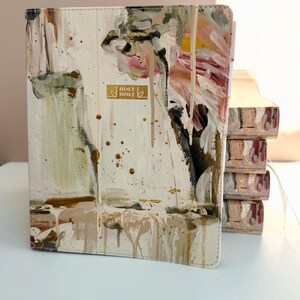 May include: A cream-colored Bible with abstract paint strokes in shades of pink, brown, and green. The cover features the gold text "HOLY BIBLE". The edges of the pages are also painted.