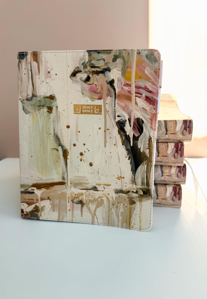 May include: A cream-colored Bible with abstract paint strokes in shades of pink, brown, and green. The cover features the gold text "HOLY BIBLE". The edges of the pages are also painted.