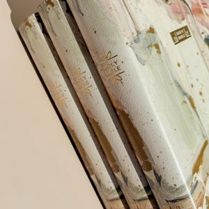 May include: Three Holy Bibles with off-white covers featuring an abstract watercolor floral design in shades of pink, green, and gold. Each spine is embossed with gold foil text reading Holy Bible and decorative flourishes, with NIV printed at the base.