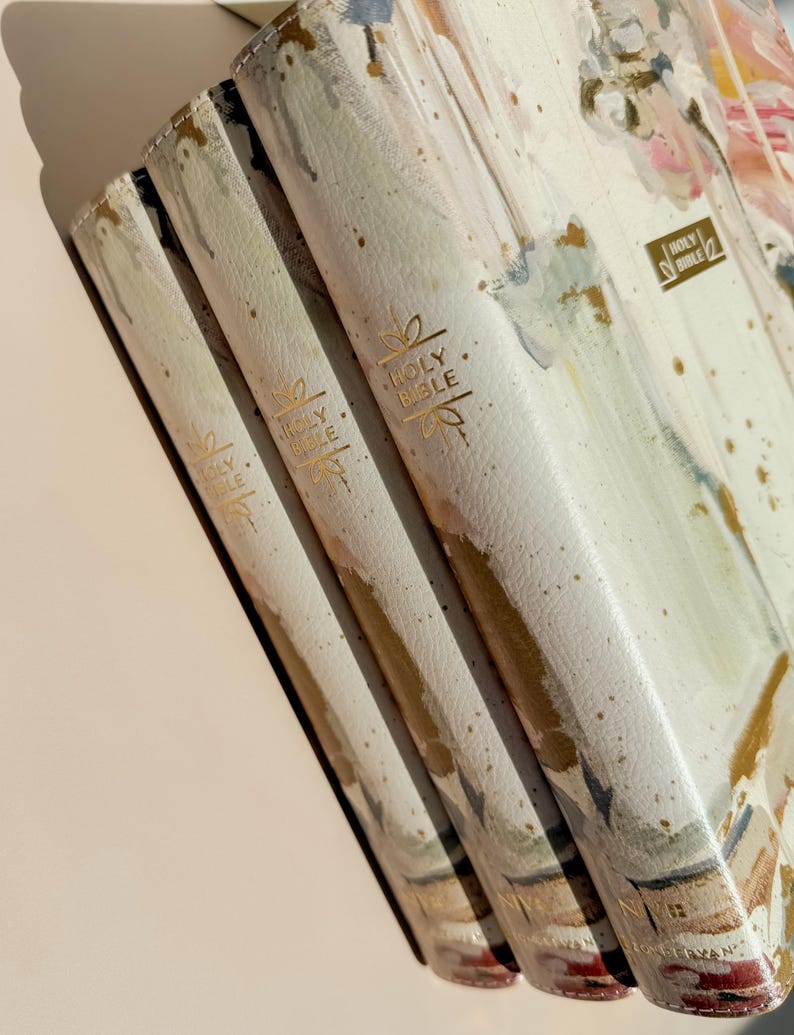May include: Three Holy Bibles with off-white covers featuring an abstract watercolor floral design in shades of pink, green, and gold. Each spine is embossed with gold foil text reading Holy Bible and decorative flourishes, with NIV printed at the base.