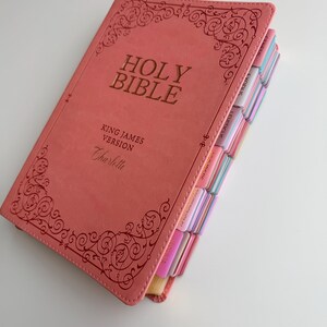 May include: A coral-colored Holy Bible in the King James Version with the name Charlotte embossed on the front cover. The cover features decorative scrollwork borders and gold lettering. The side edges include colorful index tabs for books.