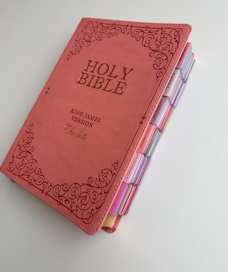 May include: A coral-colored Holy Bible in the King James Version with the name Charlotte embossed on the front cover. The cover features decorative scrollwork borders and gold lettering. The side edges include colorful index tabs for books.