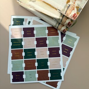 May include: A stack of books with decorative covers rests above a collection of Bible tabs. The tabs are rectangular with gold lettering and feature various book names in shades of green, brown, and pink.