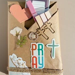 May include: A brown paper envelope containing stationery items including a peach heart cutout, a pink highlighter, pastel sticky notes, two gold cross-shaped paper clips, a small flower charm, and stickers featuring floral designs and the word PRAY.