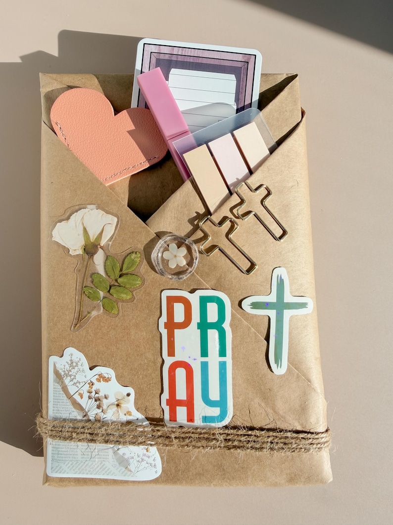 May include: A brown paper envelope containing stationery items including a peach heart cutout, a pink highlighter, pastel sticky notes, two gold cross-shaped paper clips, a small flower charm, and stickers featuring floral designs and the word PRAY.