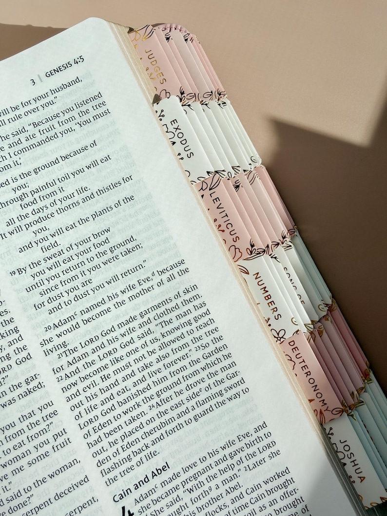 May include: An open Bible with printed text showing Genesis 4:5, featuring a series of adhesive index tabs along the edge. The tabs are alternating shades of pale pink and white, decorated with gold-colored line art of floral and leaf patterns.